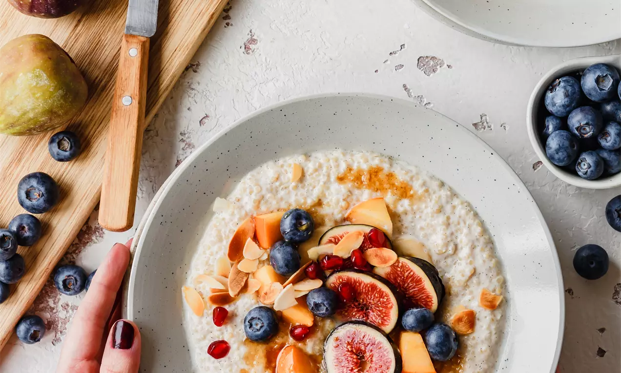 Picture - Porridge cremoso color vaniglia con fichi rosa acceso, mirtilli blu, melone arancio e mandorle su un fondo bianco punteggiato di frutti