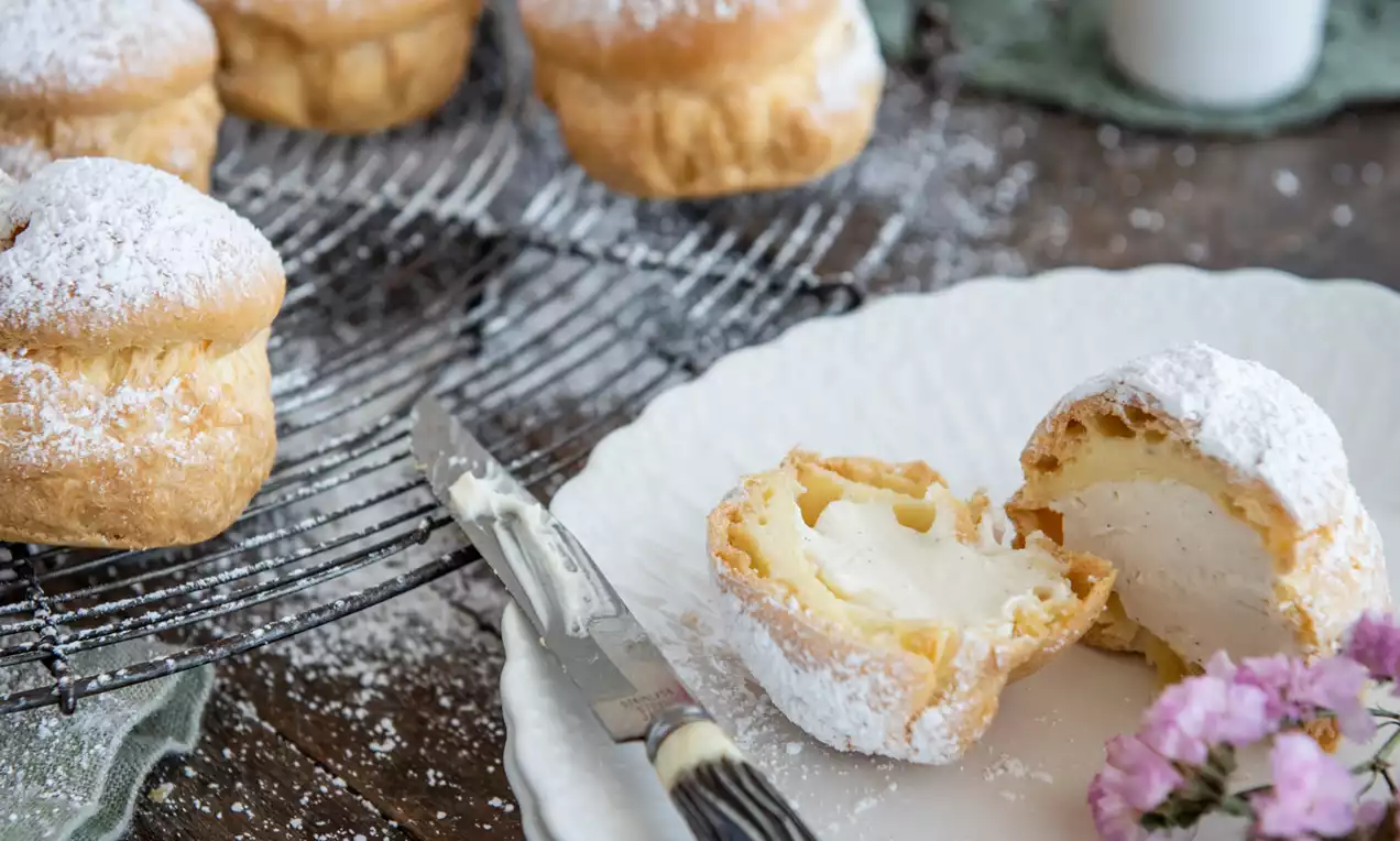 Picture - Profiteroles dorati e soffici, ripieni di crema chiara, disposti su una griglia e un piatto bianco con zucchero a velo e fiori rosa