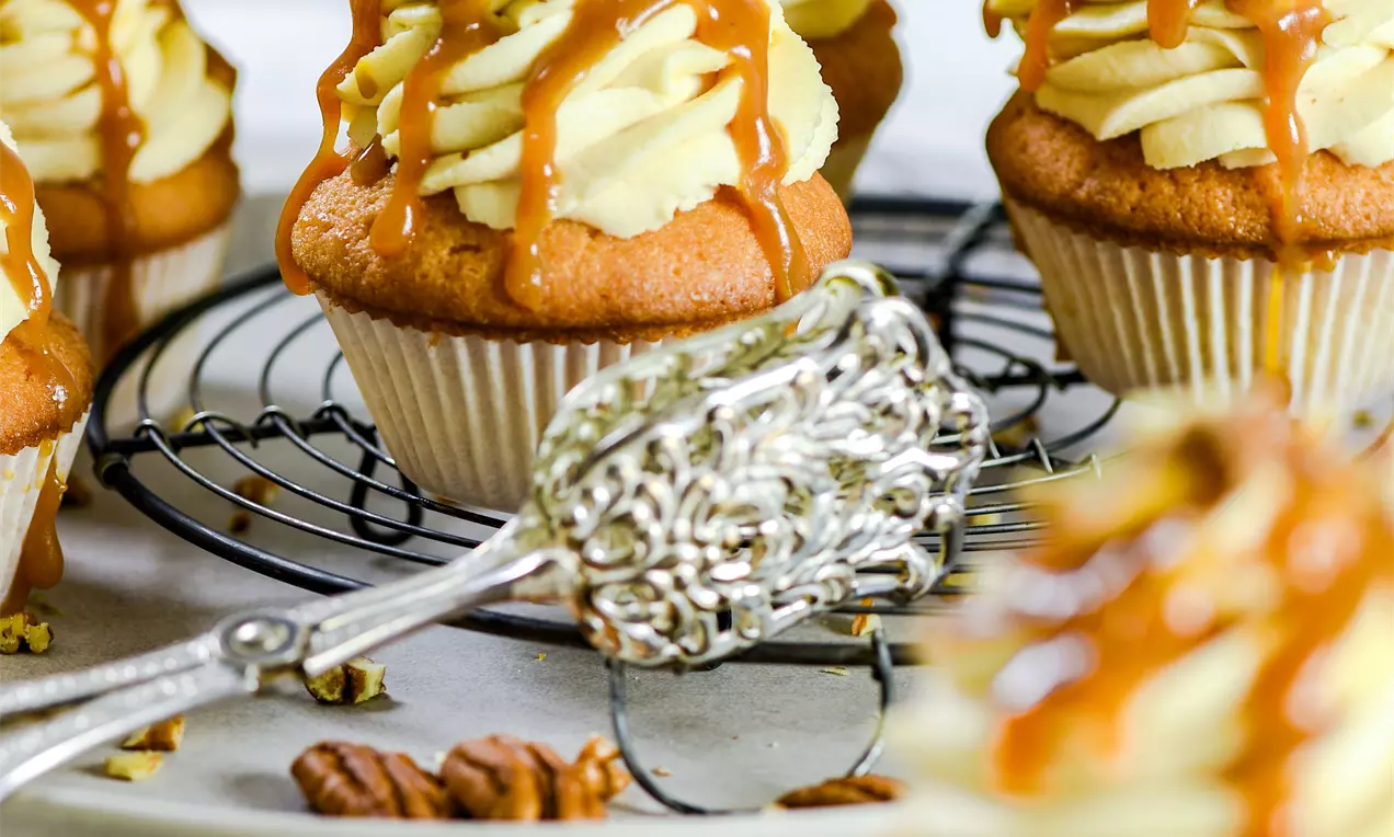 Picture - Cupcake dorati con soffice copertura di crema chiara, fili di caramello e granella di pecan su un piatto con una griglia in metallo