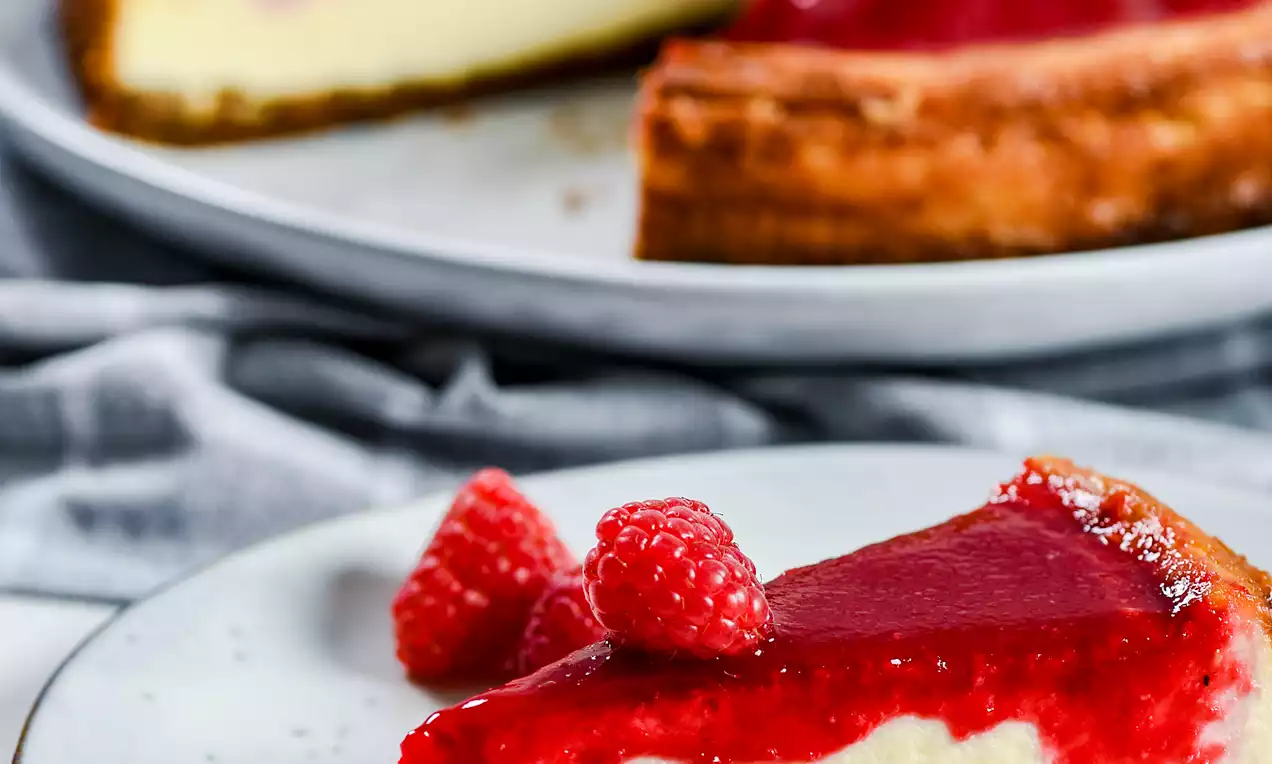 Picture - Cheesecake su base dorata di biscotto con strato cremoso e copertura lucida rosso lampone, decorata con lamponi freschi su un piatto bianco