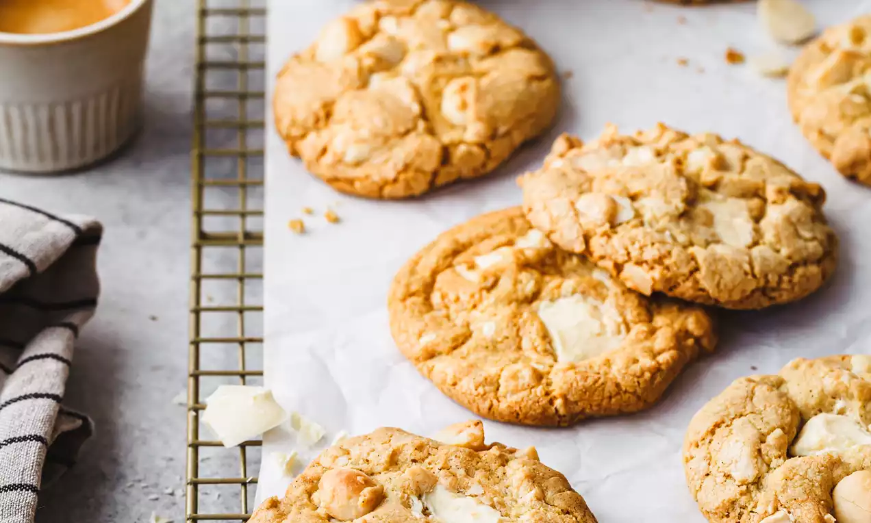 Picture - Biscotti rotondi dorati con pezzi di cioccolato bianco e noci macadamia su una griglia, circondati da tazze e ciotole su sfondo neutro