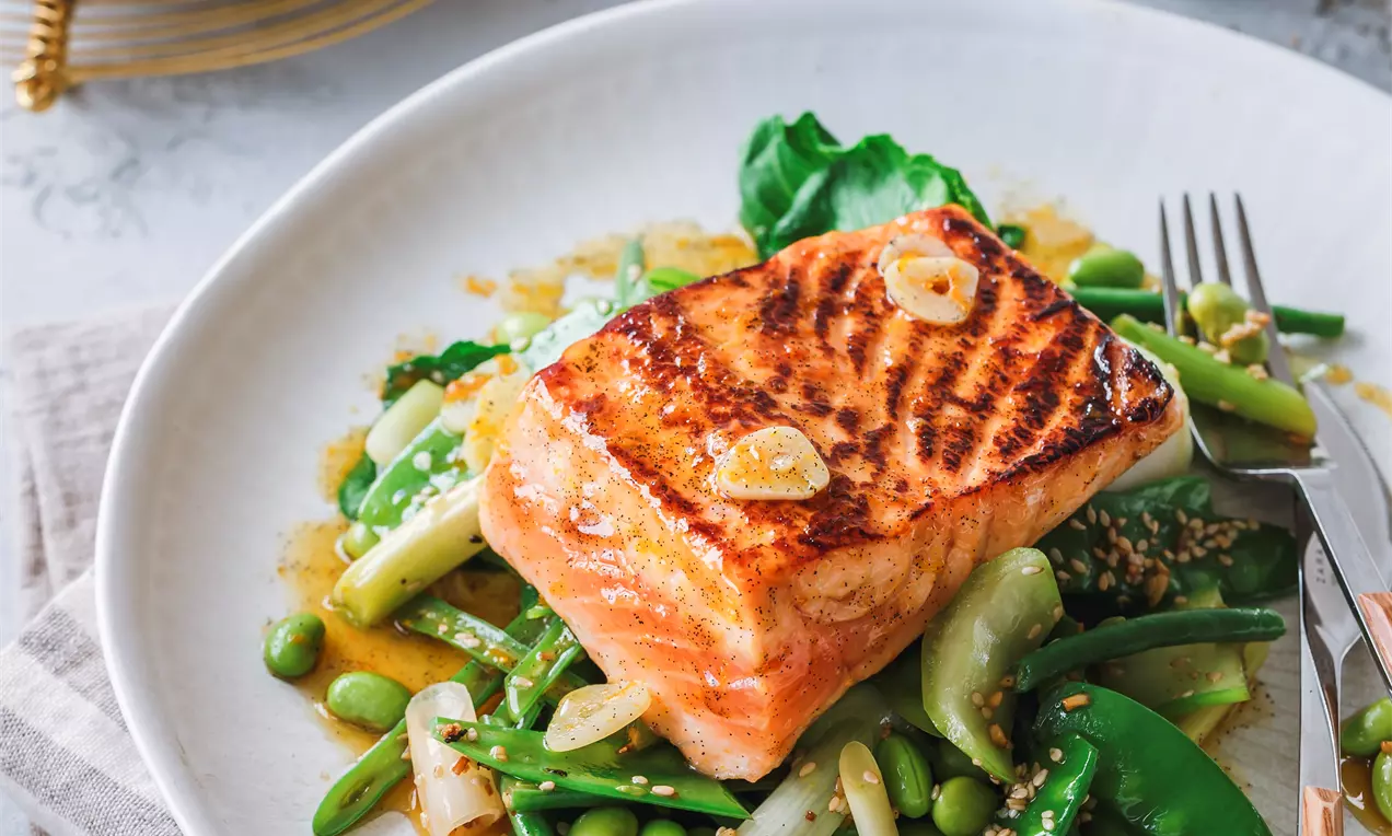 Picture - Filetto di salmone dorato con segni di griglia su letto di verdure verdi croccanti, punte di asparagi e semi di sesamo su un piatto bianco
