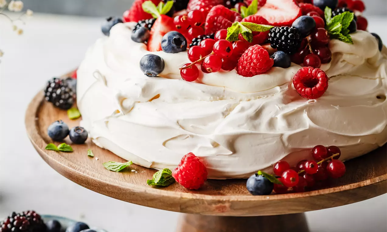 Picture - Pavlova candida e vaporosa con superficie ondulata, decorata con frutti di bosco vivaci, foglie di menta e disposta su un'alzata di legno