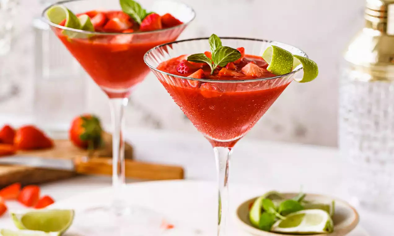 Picture - Due bicchieri da cocktail colmi di daiquiri rosso intenso, decorati con foglie di menta, lime a spicchi e fette di fragola fresca su un tavolo chiaro